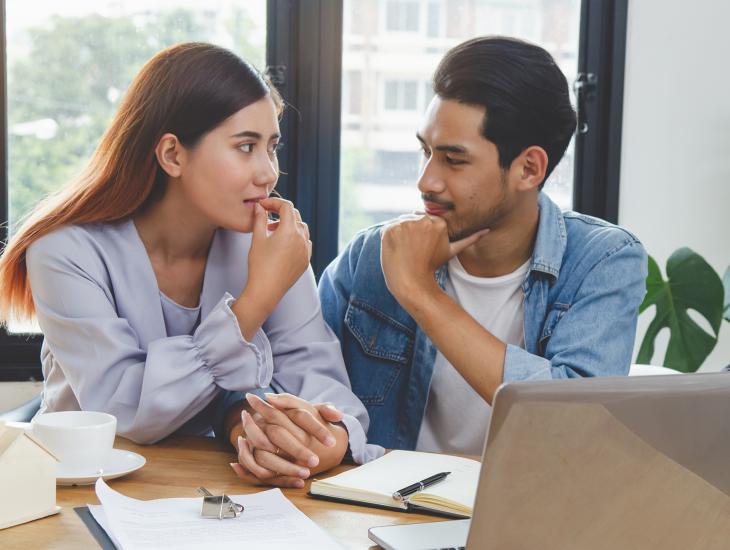 Jong stel zit aan tafel met een stapel papieren en een laptop, ze kijken elkaar aan