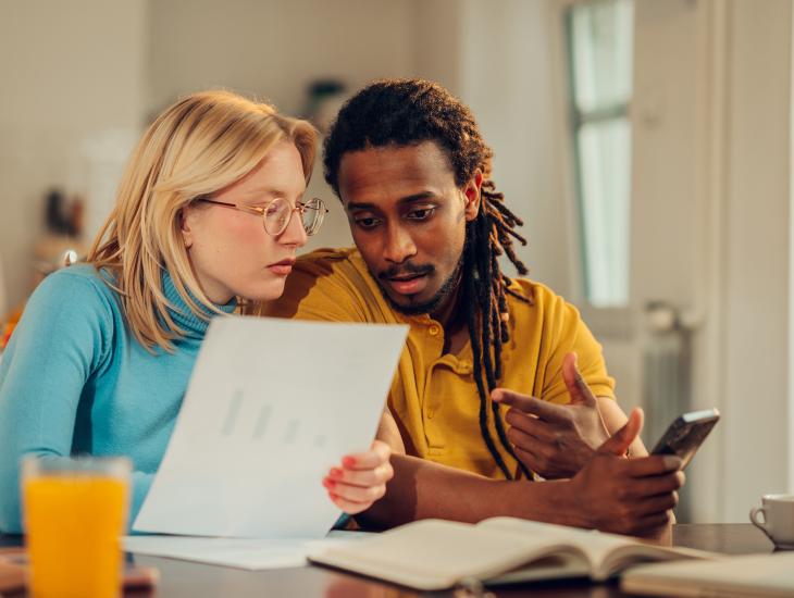 Een jong stel met zorgelijke blikken in hun ogen zit aan tafel met een stapel papieren en een rekenmachine