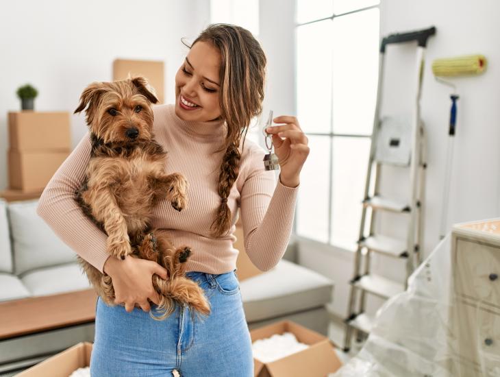 Een jonge vrouw met een hondje op haar arm staat in haar eerste koopwoning, waar ze nog aan het klussen is.