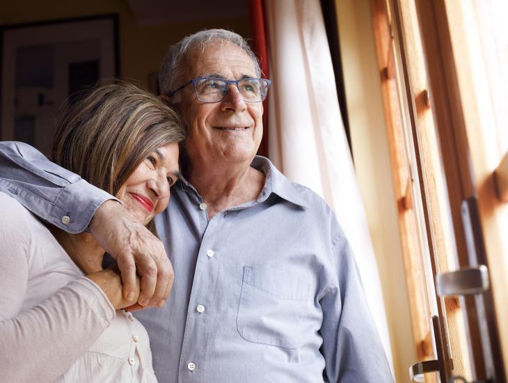Gelukkig senior stel bij het raam, haar hoofd rust op zijn schouder, hij slaat een arm om haar heen