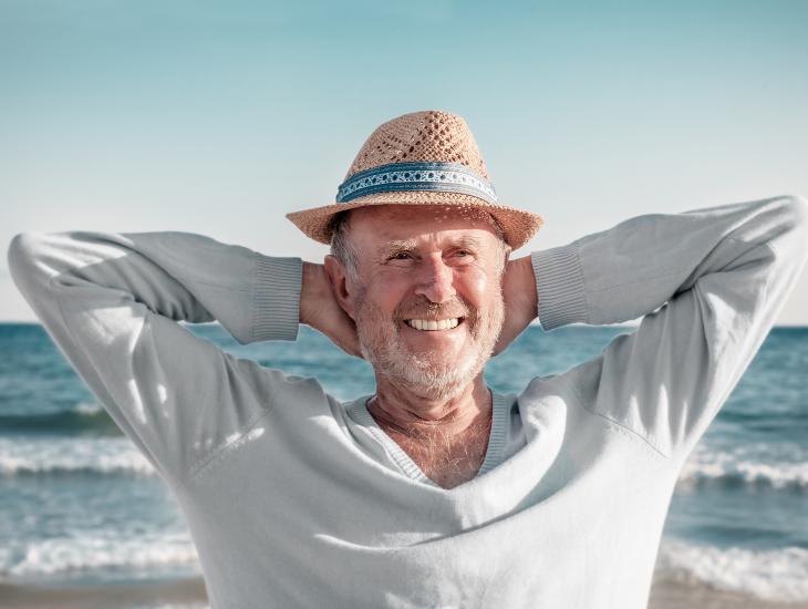 Een meneer op leeftijd zit te genieten bij de zee en vouwt zijn armen achter zijn hoofd