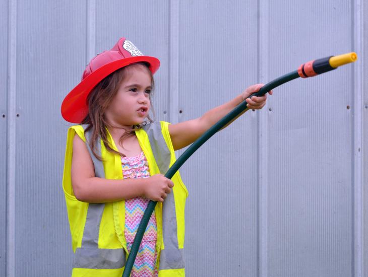 Klein meisje met helm en tuinslang speelt voor brandweervrouw
