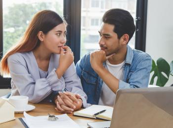Jong stel zit aan tafel met een stapel papieren en een laptop, ze kijken elkaar aan