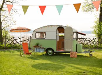 Een klein, oud caravannetje op een mooie kampeerplaats. Tussen de bomen hangen slingers.