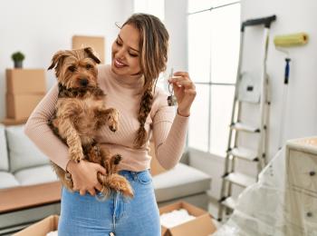 Een jonge vrouw met een hondje op haar arm staat in haar eerste koopwoning, waar ze nog aan het klussen is.