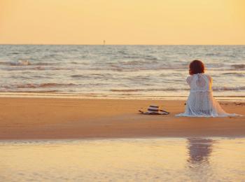 Een vrouw, op de rug gezien, zit op het strand bij zonsondergang