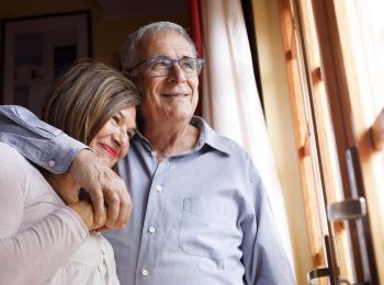 Gelukkig senior stel bij het raam, haar hoofd rust op zijn schouder, hij slaat een arm om haar heen