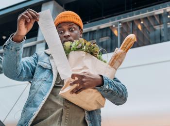 Een man met een tas vol boodschappen staart verbijsterd naar de kassabon.